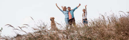 Panoramic shot of family with golden retriever waving at camera on grassy hillの写真素材