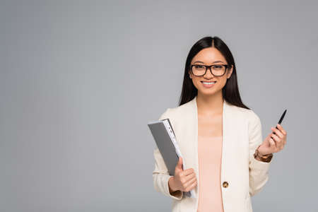 stylish asian businesswoman in white blazer and eyeglasses holding pen and folder isolated on gray