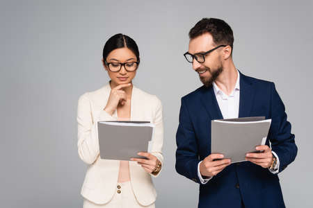 thoughtful asian businesswoman touching chin while looking at documents near colleague isolated on...
