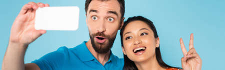 panoramic concept of asian woman showing peace gesture near boyfriend grimacing while taking selfie...