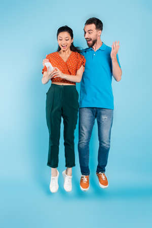 excited man touching asian woman using smartphone while levitating on blue