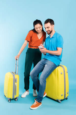 young man in polo t-shirt using smartphone while sitting on suitcase near asian woman in red blouse...