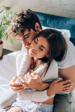 Young couple sitting in pajamas on bed and embracing each other with closed eyes