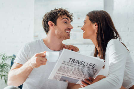 Selective focus of man holding cup of coffee and newspaper while looking at girlfriend at homeの写真素材