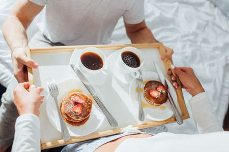 High angle view of woman holding pancakes and cups of coffee on breakfast tray near boyfriend on bedの写真素材