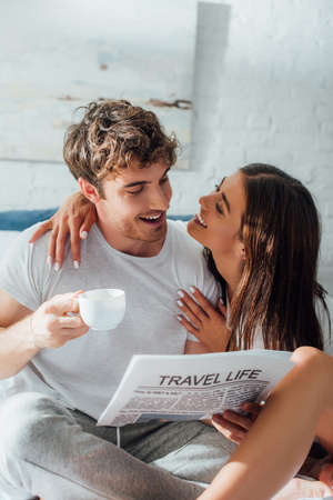 Selective focus of woman embracing and looking at boyfriend with coffee cup and newspaperの写真素材