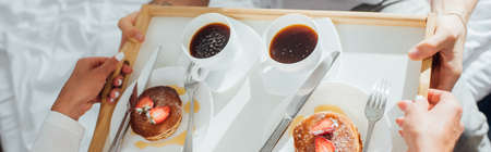 Horizontal concept of young woman holding breakfast tray with coffee and pancakes near man on bedの写真素材