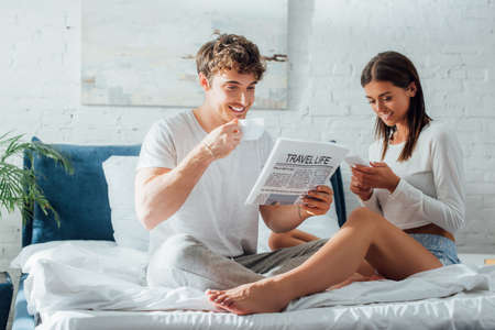 Selective focus of young couple using smartphone, reading news and holding coffee on bedの写真素材