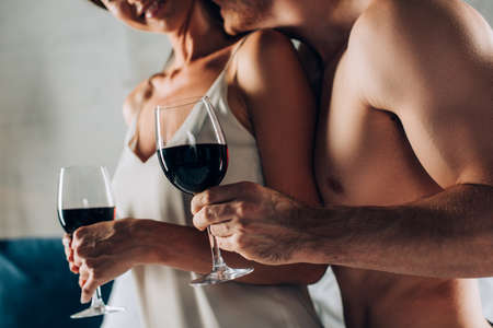Cropped view of muscular man holding glass of wine and kissing seductive woman at homeの写真素材