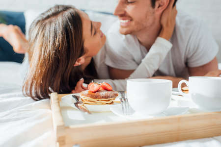 Selective focus of cups of coffee and pancakes on breakfast tray near young couple on bedの写真素材