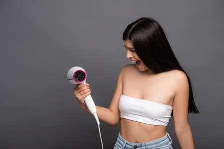brunette woman looking at hair dryer and yelling isolated on black