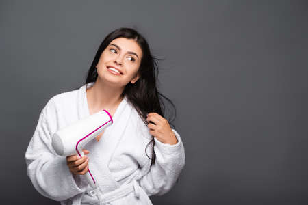 brunette long haired woman in bathrobe using hairdryer isolated on blackの写真素材
