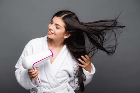 brunette long haired woman in bathrobe using hairdryer isolated on blackの写真素材