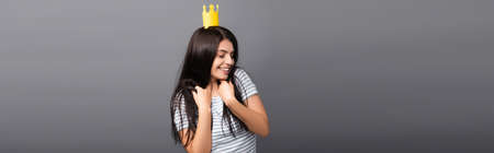 brunette long haired woman in paper crown with closed eyes smiling isolated on black, panoramic shotの写真素材