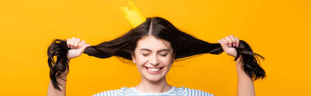 brunette woman in paper crown with closed eyes holding hair and smiling isolated on yellow, panoramic shotの写真素材