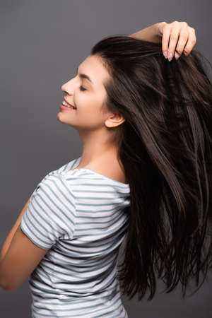 side view of brunette woman with closed eyes smiling and touching hair isolated on blackの写真素材