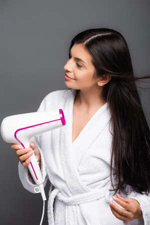 brunette long haired woman in bathrobe using hairdryer isolated on blackの写真素材