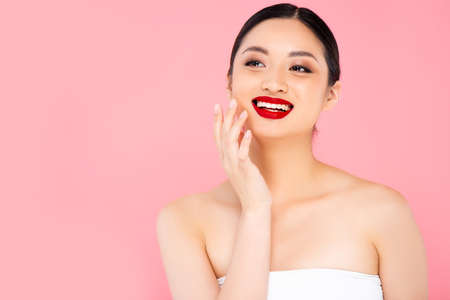 young asian woman with red lipstick on lips touching face while looking away isolated on pinkの写真素材