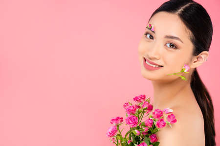 naked asian woman with floral decoration on face and petals on shoulder holding tiny roses isolated on pinkの写真素材