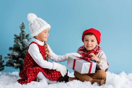 kids in hats and winter outfit sitting on snow and touching present isolated on blueの写真素材