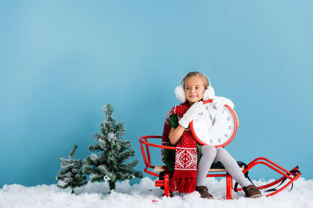 kid in winter earmuffs and scarf sitting in sleigh and holding clock near pines on blue