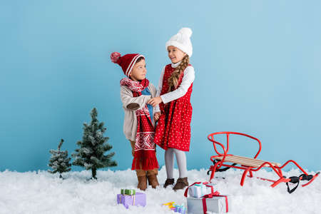 boy in hat holding hands with sister near pines, presents and sleigh isolated on blue