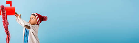 horizontal crop of boy in hat and winter outfit reaching red mailbox on blueの写真素材