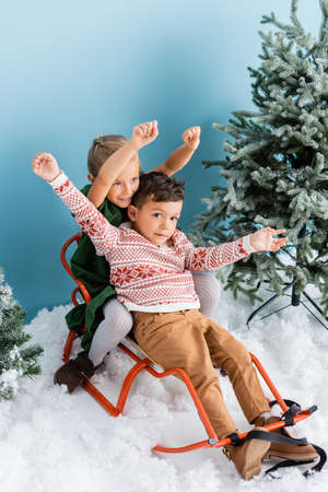 excited children with hands above heads sitting in sleigh on white snow near green pines on blueの写真素材