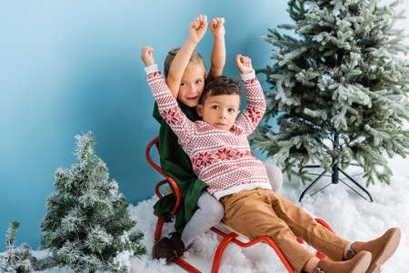 excited kids with hands above heads sitting in sleigh on white snow near green pines on blueの写真素材