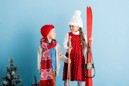 girl in hat looking at boy while holding ski poles and skis on blueの写真素材