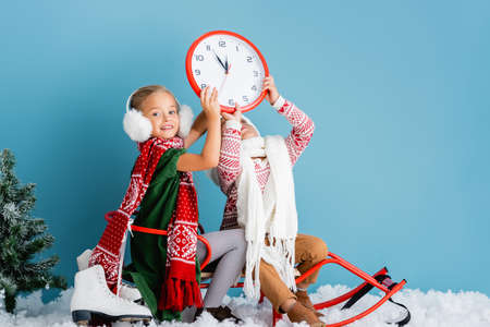 kids in winter outfit sitting on sleigh and holding clock near pine and ice skates on blueの写真素材