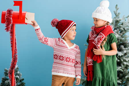 boy in knitted sweater putting envelope in mailbox and looking at sister in winter outfit isolated on blueの写真素材