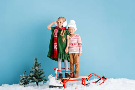 girl in winter earmuffs looking away while standing on sleigh near brother in hat and ice skates on blueの写真素材