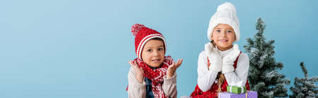panoramic shot of children in winter outfit gesturing near presents and fir isolated on blueの写真素材