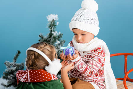 selective focus of boy in knitted hat holding present while sitting on sleight near girl in winter earmuffs on blueの写真素材
