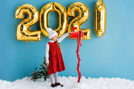 girl in hat taking present from mailbox near balloons with numbers while standing on snow on blueの写真素材
