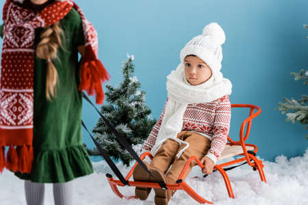selective focus of boy in winter outfit sitting on sleigh near sister on blueの写真素材