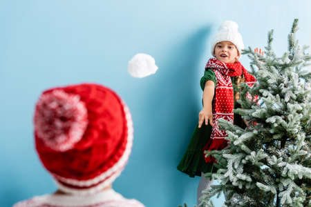 selective focus of girl in hat and scarf throwing snowball near brother on blueの写真素材