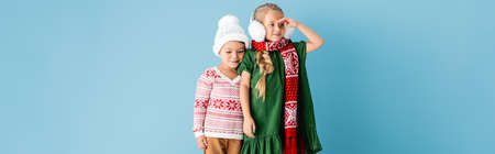 panoramic shot of girl in winter earmuffs looking away while standing near brother in hat on blueの写真素材