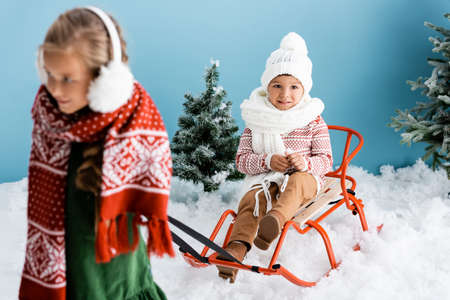 selective focus of boy in winter hat sitting on sleigh near sister on blue
