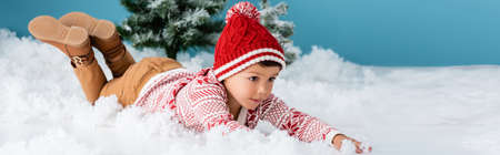 panoramic concept of boy in winter outfit lying on white snow near christmas trees on blueの写真素材