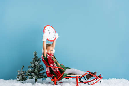 girl in winter earmuffs and scarf sitting in sleigh and holding clock above head on blueの写真素材