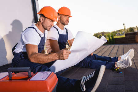 Selective focus of builders with blueprint sitting near toolbox while working on roof of house