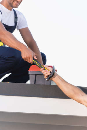 Cropped view of builder giving hammer to colleague near toolbox on roof of building isolated on...