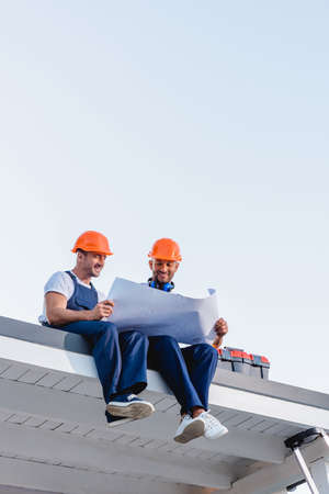 Low angle view of workmen holding blueprint while sitting near toolbox on roof of building