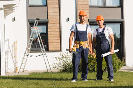 Builders in workwear holding spirit level and blueprint on lawn near building
