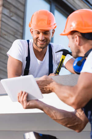 Selective focus of builder holding hammer near colleague with digital tablet on roof of building