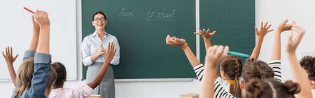 back view of multicultural pupils with hands in air, and teacher standing at chalkboard with back to school inscription, horizontal imageの写真素材