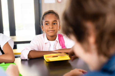 selective focus of african american schoolgirl sitting in school eatery near lunch boxの写真素材