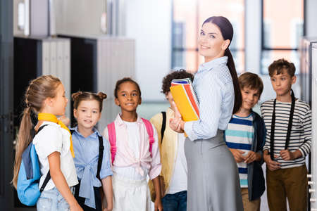 teacher looking at camera while standing near multicultural pupils in school corridorの写真素材